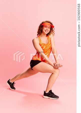 Full-length portrait of young girl in sportswear training, doing exercises, stretching against pink studio background 102900568