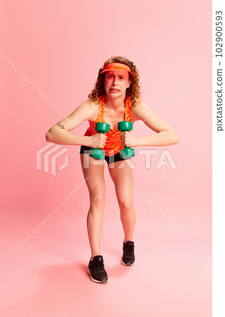 Full-length portrait of young girl in sportswear training with dumbbells with concentrated face against pink studio background 102900593