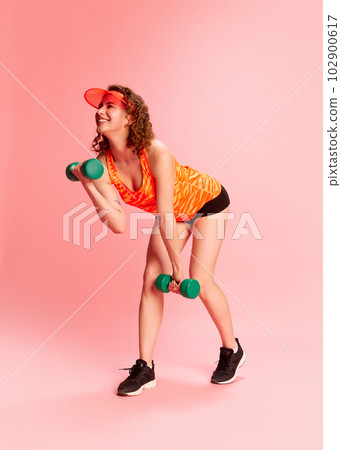 Full-length portrait of young girl in sportswear training with dumbbells, doing exercises against pink studio background Full-length portrait of young girl in sportswear training with dumbbells, doing exercises against pink studio background 102900617