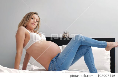 Pregnant woman sitting on a sofa at home, wearing white crop top and jeans 102907156