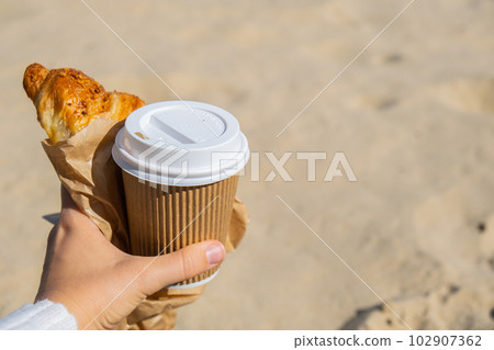 Tasty fresh croissants with coffee in paper cup in female hands on beach sea background. Breakfast outdoors wellbeing concept. FPV. Take away food Tasty fresh croissants with coffee in paper cup in female hands on beach sea background. Breakfast outdoors wellbeing concept. FPV. Take away food 102907362