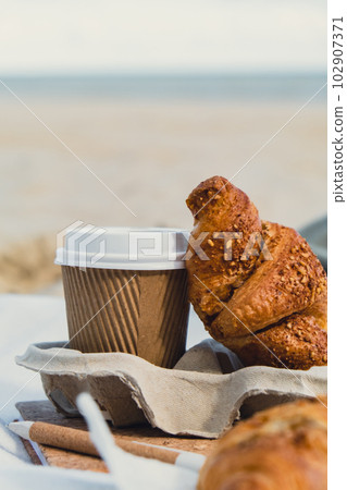 Tasty fresh croissants with coffee in paper cup on sandy beach sea coast background. Breakfast outdoors wellbeing concept. Take away food. Coffee alternatives Tasty fresh croissants with coffee in paper cup on sandy beach sea coast background. Breakfast outdoors wellbeing concept. Take away food. Coffee alternatives 102907371