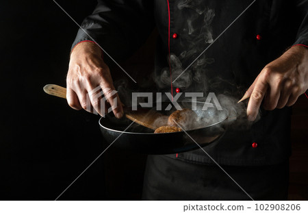 The chef fries meat patties for a burger in a frying pan. Space for hotel menu on black background. The chef fries meat patties for a burger in a frying pan. Space for hotel menu on black background. 102908206