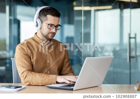 A young man sits at a workplace in the office, works, studies on a laptop with headphones. Listens to the webinar, music, course. A young man sits at a workplace in the office, works, studies on a laptop with headphones. Listens to the webinar, music, course. 102908887