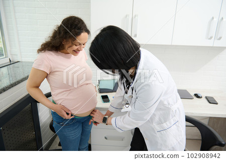 The female doctor obstetrician gynecologist measures the belly of a pregnant woman with a measuring tape, during regular medical check-up in the pregnancy. Gynecological examination and consultation The female doctor obstetrician gynecologist measures the belly of a pregnant woman with a measuring tape, during regular medical check-up in the pregnancy. Gynecological examination and consultation 102908942