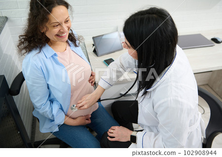 Doctor gynecologist listening to pregnant patient's belly. Smiling happy gravid female expectant mother visits obstetric clinic for regular checkup during pregnancy. Gynecology. Women's health concept Doctor gynecologist listening to pregnant patient's belly. Smiling happy gravid female expectant mother visits obstetric clinic for regular checkup during pregnancy. Gynecology. Women's health concept 102908944