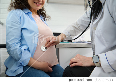 Close-up of doctor obstetrician gynecologist putting stethoscope on pregnant woman belly and listening the heartbeat of future newborn baby, examining the gravid mother in medical gynecological clinic 102908945