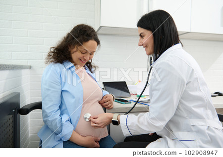 Confident female doctor obstetrician gynecologist checking heartbeat of the baby in womb using a stethoscope, examining a pregnant woman in gynecological clinic. Gynecology consultation. Pregnancy Confident female doctor obstetrician gynecologist checking heartbeat of the baby in womb using a stethoscope, examining a pregnant woman in gynecological clinic. Gynecology consultation. Pregnancy 102908947