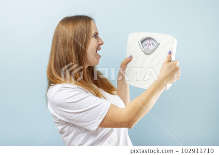 Young brunette woman looking at weight scale in shock on blue background Young brunette woman looking at weight scale in shock on blue background 102911710