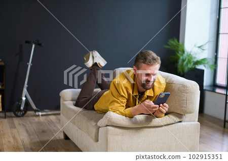 Smiling man enjoying time at home, sitting on sofa with phone. He appears handsome and content engages in texting or messaging on mobile device. High quality photo Smiling man enjoying time at home, sitting on sofa with phone. He appears handsome and content engages in texting or messaging on mobile device. High quality photo 102915351