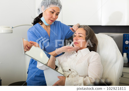 Young female cosmetologist attentive examines the face of an elderly female client before the procedure 102921811