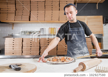 The chef prepares pizza. Raw pizza ready to bake. Cook in a blue apron in the kitchen. with a shovel in his hands. boxes for food delivery on background. The chef prepares pizza. Raw pizza ready to bake. Cook in a blue apron in the kitchen. with a shovel in his hands. boxes for food delivery on background. 102927580
