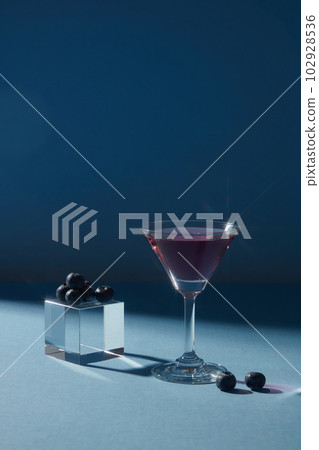 Transparent cube with blueberries placed on, decorated with a cocktail glass of purple liquid over a dark background. Promoting product extracted from Blueberry (Vaccinium Corymbosum) 102928536