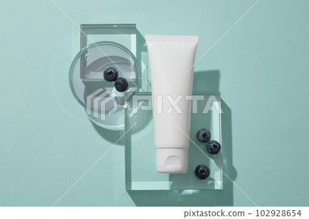 Top view of cosmetics tube mockup for design placed on transparent podiums, blueberries and essence on petri dish display on blue background. Scene for promote cosmetic, copy space 102928654