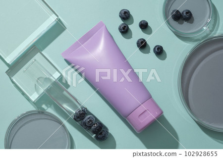 Mockup scene with purple bottle for cosmetics of blueberry extract on blue background. Petri dish filled essence and test tube decorated with fresh blueberries. Space for design 102928655