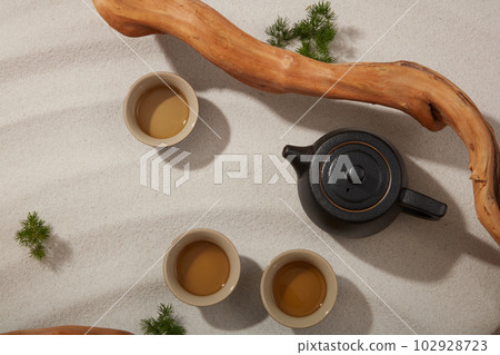 Top view of black teapot and tea set decorated with dried twig and green grass on beige sand background. Minimal background for product presentation 102928723