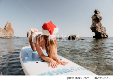 Woman sea sup. Close up portrait of happy young caucasian woman with long hair in Santa hat looking at camera and smiling. Cute woman portrait in a white bikini posing on sup board in the sea Woman sea sup. Close up portrait of happy young caucasian woman with long hair in Santa hat looking at camera and smiling. Cute woman portrait in a white bikini posing on sup board in the sea 102930839