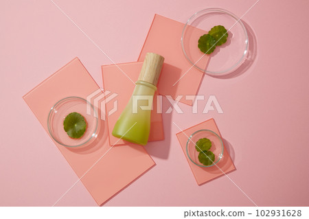 A green glass bottle placed on some acrylic sheets decorated on pink background with gotu kola leaves in petri dishes. Minimal scene for mockup packaging, cosmetics design. A green glass bottle placed on some acrylic sheets decorated on pink background with gotu kola leaves in petri dishes. Minimal scene for mockup packaging, cosmetics design. 102931628
