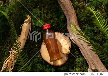 Natural concept for advertising product with herbal medicine. A glass bottle unlabeled on wooden podium, ginseng root and twigs on moss background. Space for design. 102931875