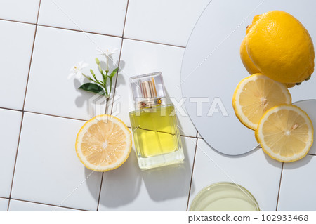 Against a white mosaic tiles background, a perfume transparent bottle displayed with flower branch, mirror and a petri dish of yellow liquid. Lemon (Citrus limon) essential oil creates a feeling of co 102933468