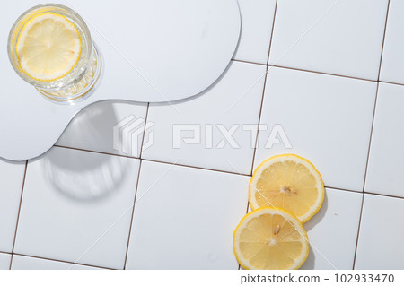 A geometric mirror with a glass of Lemon slice placed on against a white mosaic tiles background. Lemon (Citrus limon) contains a lot of potassium to help stimulate brain activity. Empty space 102933470