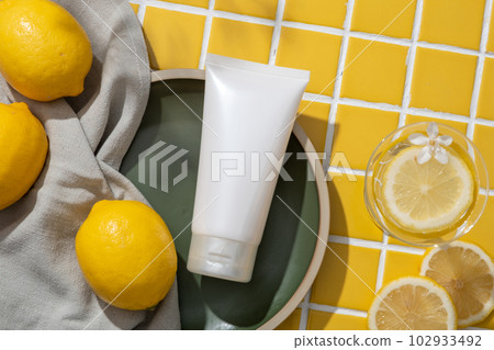 Top view of white cosmetic container and a towel placed on a dish displayed with a glass of Lemon slice and white flower. Lemon (Citrus limon) can get rid of dead skin cells 102933492