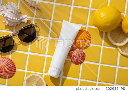 Flat lay of an unbranded cosmetic container decorated with Lemons, a sunglasses and colorful seashells. Lemon (Citrus limon) is a natural method of increasing collagen in facial skin 102933498