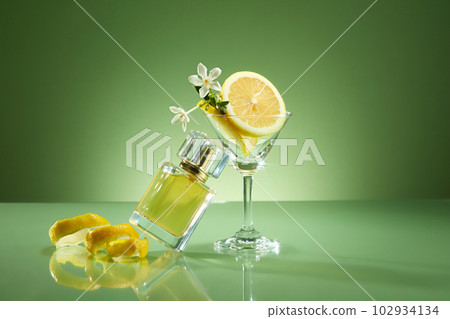 Perfume bottle prototype without label, slice and peel of lemon and glass cup decorated on green background. Fragrance oil from lemon essential oil has a fresh, refreshing, pleasant scent 102934134