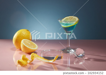 Peel and slices of fresh lemon are decorated on dark blue background with glass cup and crystal ball. Empty space on transparent podium for display products of lemon extract. Advertising photo. Peel and slices of fresh lemon are decorated on dark blue background with glass cup and crystal ball. Empty space on transparent podium for display products of lemon extract. Advertising photo. 102934152