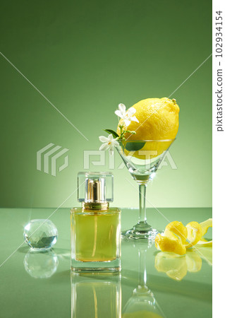 Scene for advertising fragrance oil products from lemon oil. Perfume bottle prototype unlabeled with glass cup containing lemon and white flower, crystal ball and peel of lemon on green background 102934154