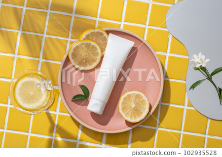 Some Lemon slices and a white tube put on a pink dish. A mirror, white flowers and a glass are displayed. Shadow of tropical leaves. Lemon (Citrus limon) is a popular fruit for its vitamin C content 102935528