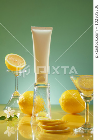 Blank label white tube standing on an upside down glass, arranged with several Lemon slices, flowers and glasses. Lemon (Citrus limon) is considered a vegan ingredient 102935536