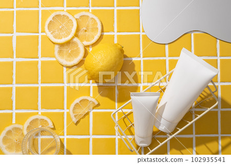 Two cosmetic tubes inside a basket, decorated with several Lemon slices, a glass of water and a mirror. Lemon (Citrus limon) has astringent qualities due to its acidic level 102935541