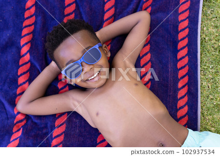 High angle view of african american smiling shirtless boy wearing sunglasses and lying on towel 102937534
