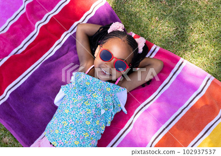 Cute african american girl wearing sunglasses with hands behind head lying on towel 102937537