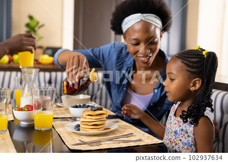 African american mother pouring honey on pancakes while having breakfast with daughter at home 102937634