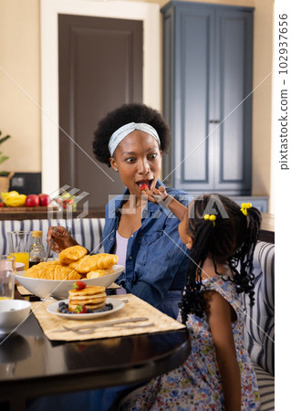 African american girl feeding strawberry to mother while having breakfast at dining table at home 102937656