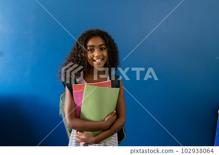 Portrait of happy biracial schoolgirl with school bag holding notebooks on blue background Portrait of happy biracial schoolgirl with school bag holding notebooks on blue background 102938064