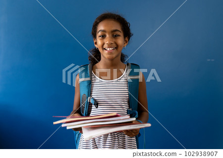 Portrait of happy biracial schoolgirl holding notebooks smiling, on blue background with copy space 102938907