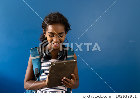 Happy biracial schoolgirl with headphones using tablet, smiling on blue background with copy space Happy biracial schoolgirl with headphones using tablet, smiling on blue background with copy space 102938911