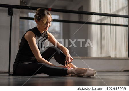 An Asian ballerina sits on the floor and puts on pointe shoes. 102938939