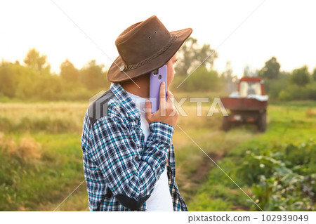 Farmer talking on the purple phone on the tractor background. Amidst the vast expanse of a field, a farmer confidently walks, seamlessly multitasking by engaging in a conversation on mobile phone 102939049