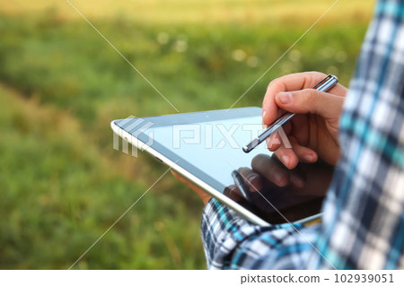 Tablet screen and stylus. An agronomist farmer man is seen using a digital tablet computer amidst a young cornfield during the serene hours of sunrise or sunset. Close up hands 102939051