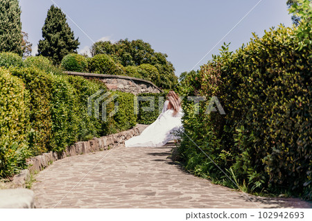 Brunette runs white dress park. A beautiful woman with long brown hair and a long white dress runs along the path along the beautiful bushes in the park, rear view Brunette runs white dress park. A beautiful woman with long brown hair and a long white dress runs along the path along the beautiful bushes in the park, rear view 102942693