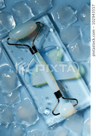 Blue jade face roller and Gua Sha scraping for beauty facial massage therapy on ice cubes and water background. 102943557