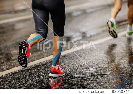 legs female runner athlete run marathon race on wet asphalt, blue kinesio tape on shins legs female runner athlete run marathon race on wet asphalt, blue kinesio tape on shins 102950845