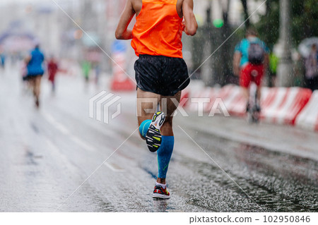rear view runner athlete run marathon race on rain, in blue compression sleeve on feet and orange singlet 102950846