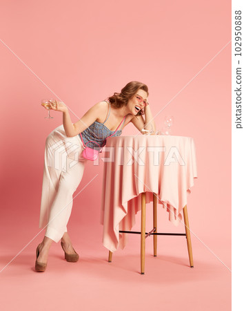 Portrait of beautiful young woman in stylish clothes standing at table with champagne glasses, drinking and laughing against pink studio background 102950888