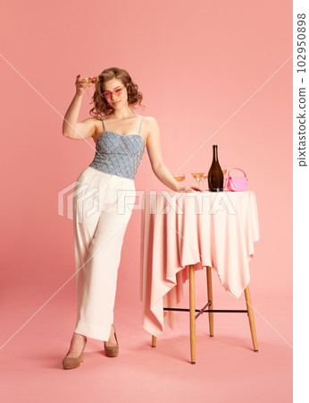 Full-length portrait of young, beautiful woman in stylish outfit at party, raising glass with champagne against pink studio background Full-length portrait of young, beautiful woman in stylish outfit at party, raising glass with champagne against pink studio background 102950898