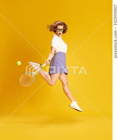 Full-length portrait of young active girl in sportswear jumping with tennis racket against yellow studio background Full-length portrait of young active girl in sportswear jumping with tennis racket against yellow studio background 102950907